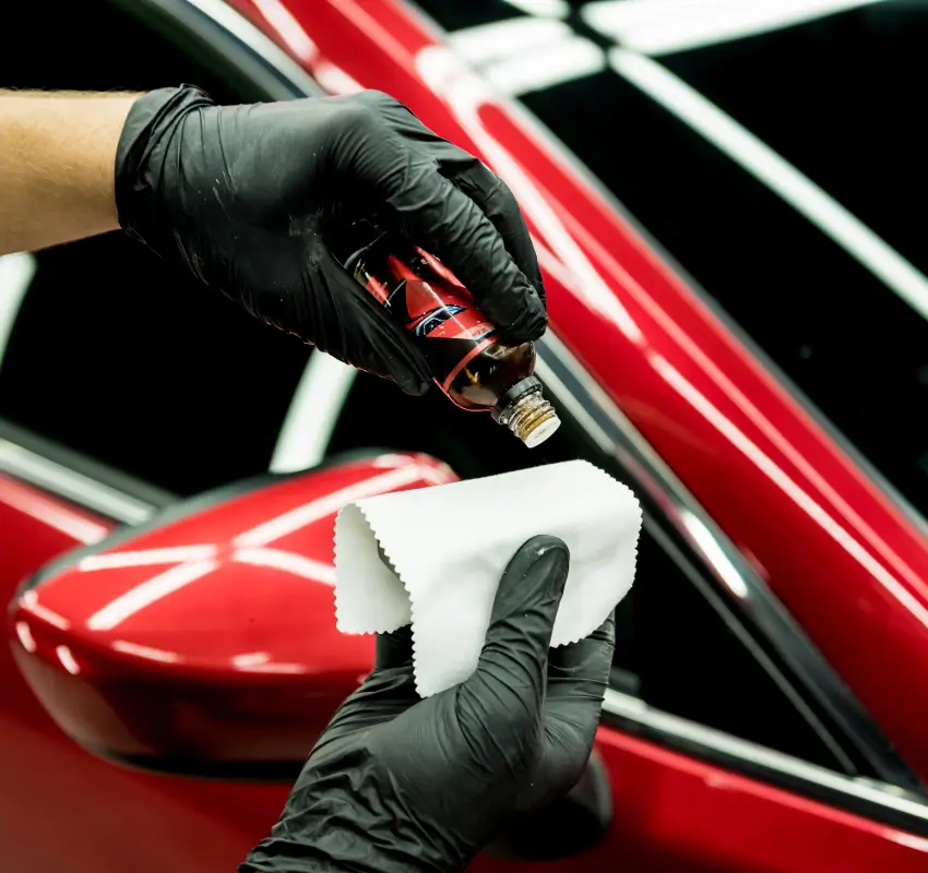 Professional auto detailer in black gloves applying ceramic coating liquid to a white applicator cloth.