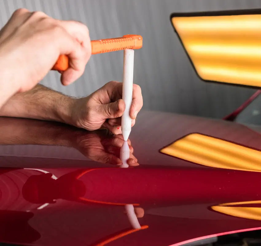 Close-up of a technician performing paintless dent repair (PDR) on a red car using a specialized tap-down tool.